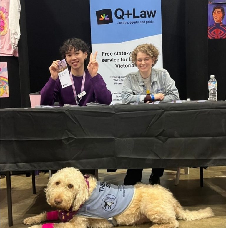 Two people sit behind a table, in front of a Q+Law banner. A fluffy dog lies in front of the table.